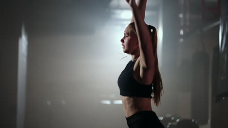 In-slow-motion-a-woman-lifts-a-dumbbell-over-her-head-while-working-out-in-a-dark-gym.-Athletic-strong-woman-performs-a-difficult-workout-with-lifting-dumbbells